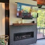 A modern living room with a wall-mounted television above a brick fireplace, a wooden mantel with decorative items, and a basket filled with logs next to it. A large window overlooks greenery.