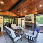 An outdoor living space with a wooden ceiling, cozy seating, a fireplace, a ceiling fan, and glass doors opening to a lush garden.