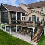 A large modern house with a two-story deck featuring outdoor seating areas. The upper deck has a screened section and a stairway leading to a tiled patio below. The house is surrounded by a well-kept lawn.