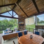 A covered patio with a wooden ceiling features a seating area, a dining table, a ceiling fan, a TV mounted on a brick wall, and a grill. The patio overlooks a grassy yard and neighboring houses.