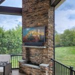 Outdoor patio with a stone fireplace featuring a mounted TV, surrounded by greenery. The ceiling is wooden with exposed beams, and there is a section of a sofa visible in the foreground.